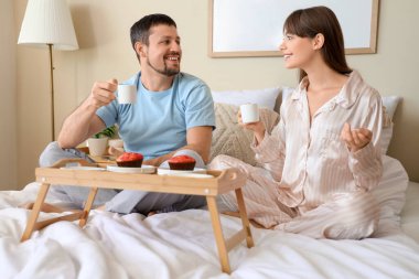 Young happy couple in pajamas with cups of coffee and cupcakes having breakfast together at home clipart