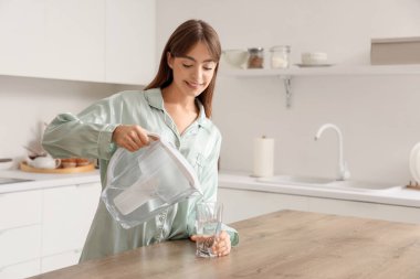 Young woman pouring water from filter jug into glass on table in kitchen clipart