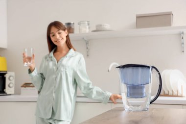 Water filter jug on table against young woman with glass in kitchen, closeup clipart