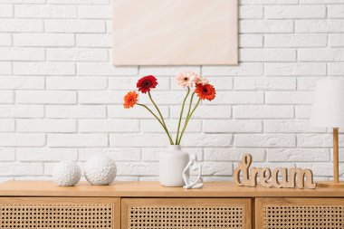 Gerbera çiçekli vazo ve beyaz tuğla duvarın yanındaki çekmecelerin üzerindeki heykel.
