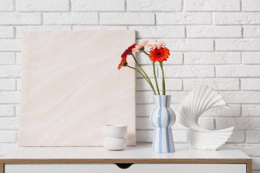 Gerbera çiçekli vazo, beyaz tuğla duvarın yanındaki çekmecelerde resim ve heykel.