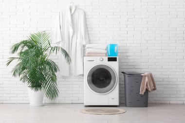 Modern çamaşır makinesi, çamaşır sepeti, ev bitkisi ve bornoz.