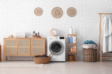 Modern çamaşır makinesi, sepetler, çamaşır deterjanı ve çekmeceli raf ünitesi.