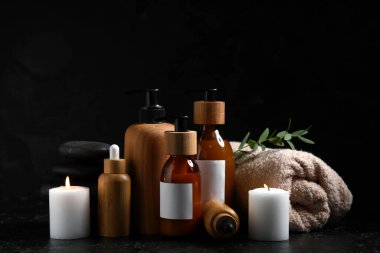 Farklı kozmetik ürünleri seti, yanan mumlar, havlu ve kaplıca taşları koyu arka planda.