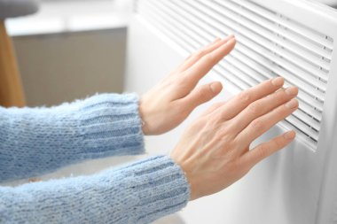 Yatak odasında elektrikli konteyner ısıtıcısının yanında ellerini ısıtan genç bir kadın var.