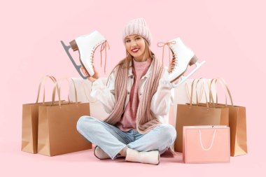 Buz patenleri ve alışveriş torbalarıyla pembe arka planda oturan, kışlık giysili güzel bir kadın.