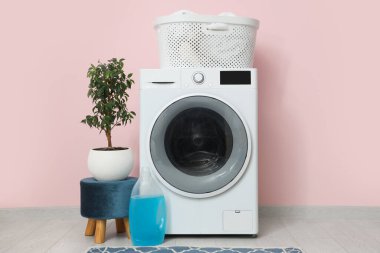 Pembe duvarın yanında sepetli, çiçekli ve deterjanlı modern çamaşır makinesi.