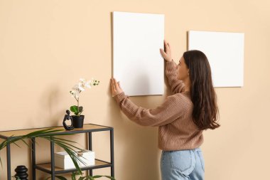 Oturma odasındaki bej duvara boş tuval asan genç bir kadın.