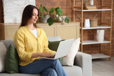 Evdeki gri koltukta dizüstü bilgisayar kullanan genç bir kadın.