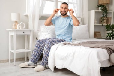 Yatak odasında oturan, kulak tıkacı takan genç adam.