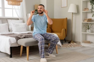 Yatak odasında oturan, kulak tıkacı takan genç adam.