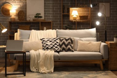 Modern oturma odasının içinde gri kanepe, sehpada dizüstü bilgisayar ve akşam lambaları.