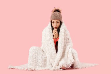 Pembe arka planda oturan, sıcak ekoseli, şapkalı, donmuş genç bir kadın.