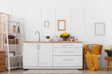 Mutfağın içinde tezgahlar, raf ünitesi ve kasımpatı çiçekleri var.