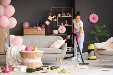 Doğum günü partisinden sonra dağınık odada elektrikli süpürge kullanan genç Asyalı kadın.