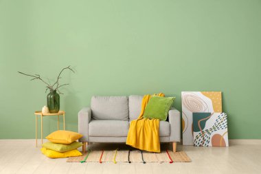 Konforlu kanepe, resimler, yastıklar ve vazo. Odadaki yeşil duvarın yanındaki sehpanın üzerinde dallar var.