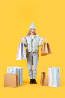 Sarı arka planda alışveriş torbaları olan, kışlık giysili güzel bir kadın.