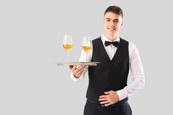 Молодой официант с бокалами вина на подносе на сером фоне