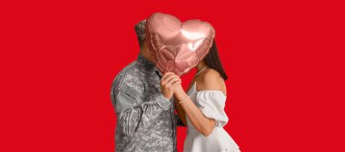 Asker ve genç kadın kırmızı arka planda kalp şeklinde bir balonun arkasında öpüşüyor. Sevgililer Günü kutlaması