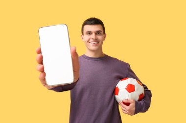 Sarı arka planda futbol topu ve cep telefonu olan genç bir adam. Spor bahsi kavramı