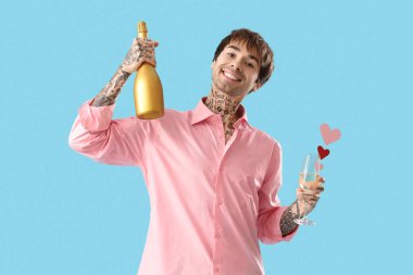 Handsome young man holding bottle and glass of champagne with paper hearts on blue background. Valentine's day celebration