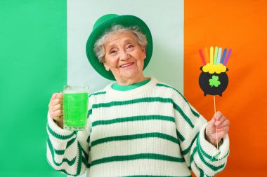 İrlanda bayrağına karşı Aziz Patrick Günü kutlamaları için yeşil bira ve dekorla cüce şapkalı mutlu bir son sınıf kadını.