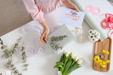 Tebrik kartı, hediye kutusu, çiçekler ve ışık masasında paketleme malzemeleri olan kadın elleri. Uluslararası Kadınlar Günü