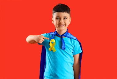 Süper kahraman gibi giyinmiş küçük çocuk kırmızı arka planda altın kurdeleyi işaret ediyor. Çocukluk kanseri farkındalığı