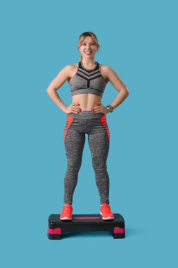 Genç bayan fitness eğitmeni mavi arka planda aerobik basamağında duruyor.