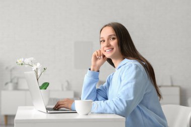 Genç, yorgun bir kadın bir fincan kahveyle masada oturuyor ve şık bir odada dizüstü bilgisayar kullanıyor.