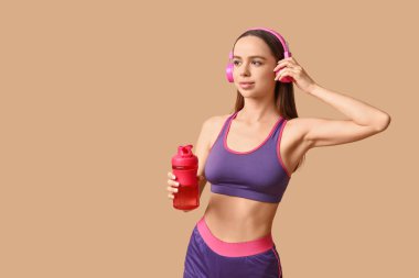 Genç bayan antrenör bej arka planda bir şişe suyla müzik dinliyor.