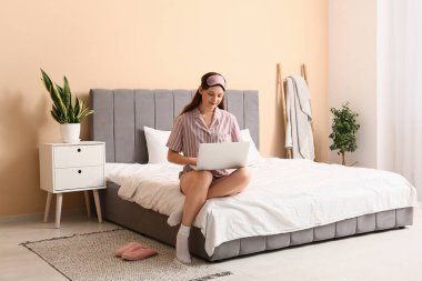 Pijamalı ve maskeli genç bir kadın evde dizüstü bilgisayar kullanıyor. Uzak çalışma konsepti