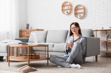 Pijamalı ve gözlüklü genç bir kadın dizüstü bilgisayarla çalışıyor ve evdeki kanepenin yanında yerde kahve içiyor. Uzak çalışma konsepti