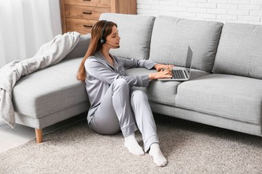 Pijamalı ve kulaklıklı genç bir kadın evdeki kanepenin yanında modern dizüstü bilgisayarla çalışıyor. Uzak çalışma konsepti
