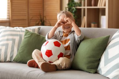 Koltuğunda oturan, elinde futbol topu olan ve elleriyle kalp yapan tatlı bir çocuk.