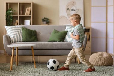 Küçük çocuk oturma odasındaki kanepenin yanında futbol topuyla oynuyor.
