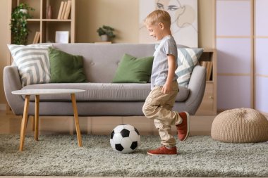Küçük çocuk oturma odasındaki kanepenin yanında futbol topuyla oynuyor.