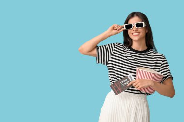 3 boyutlu sinema gözlüklü, patlamış mısır kovalı ve mavi arka planda bileti olan genç ve güzel bir kadın.