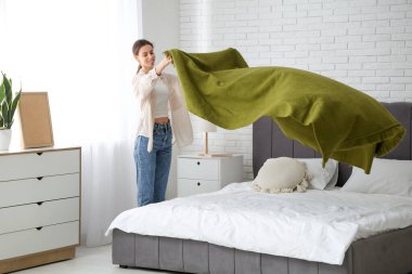 Evdeki yatağa yeşil battaniye örten genç bir kadın.