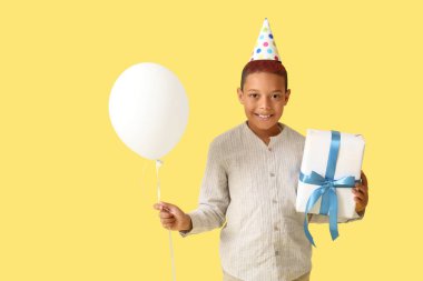 Hava balonu ve hediye kutusuyla sarı arka planda doğum gününü kutlayan parti şapkalı tatlı Afro-Amerikalı çocuk.