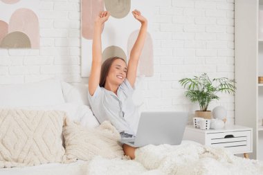 Evde dizüstü bilgisayarı olan genç bir kadın yatakta uzanıyor.