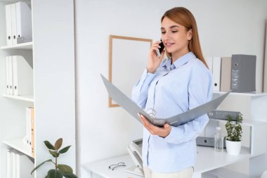 Ofiste cep telefonuyla konuşan belge klasörlü genç bir iş kadını.