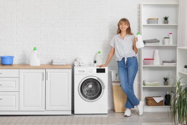 Young woman with bottle of laundry detergent near washing machine at home clipart