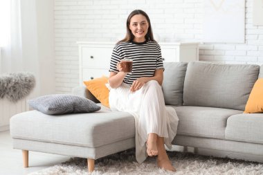 Oturma odasındaki kanepede oturan cam bardak sıcak kakaolu mutlu genç kadın.