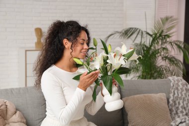 Güzel, mutlu, Afro-Amerikan bir kadın. Elinde bir buket beyaz zambak çiçeğiyle evdeki koltukta oturuyor.