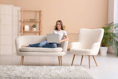 Genç mutlu kadın kanepede uzanıyor ve oturma odasında dizüstü bilgisayar kullanıyor.