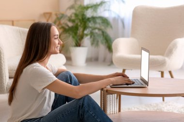 Genç, mutlu bir kadın oturma odasındaki sehpada dizüstü bilgisayar kullanıyor.