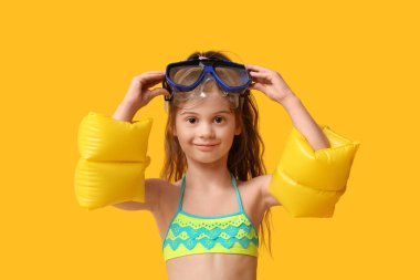 Sarı arka planda şişme kollukları ve şnorkel maskesi olan mayo giymiş şirin bir kız.