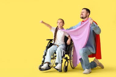 Little girl in wheelchair and her father with superhero capes on yellow background clipart