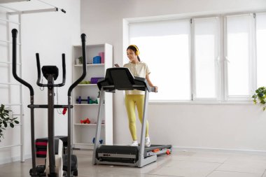 Spor salonundaki koşu bandında cep telefonu eğitimi almış kulaklıklı Asyalı bir kadın.